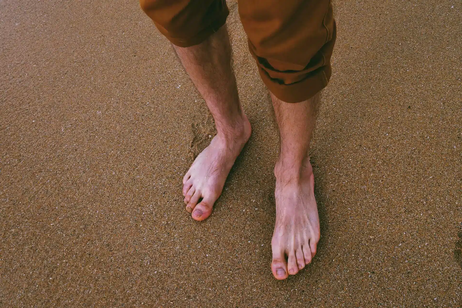 Beine einer Person mit hochgekrempelter Hose die an einem Sandstrand steht